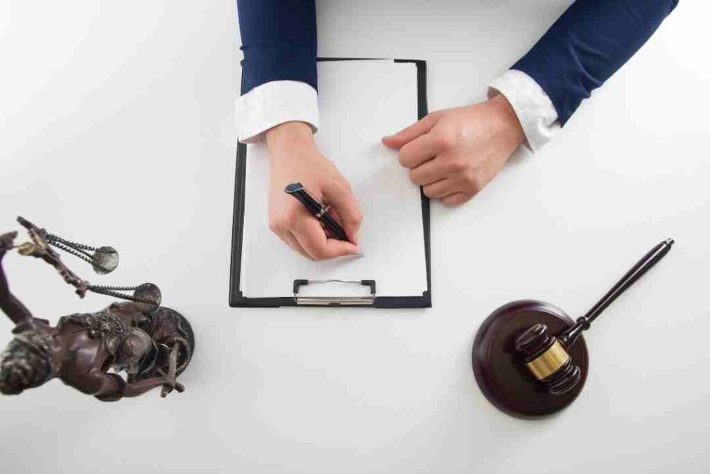 Hands writing on a clipboard with a gavel and Justice statue.